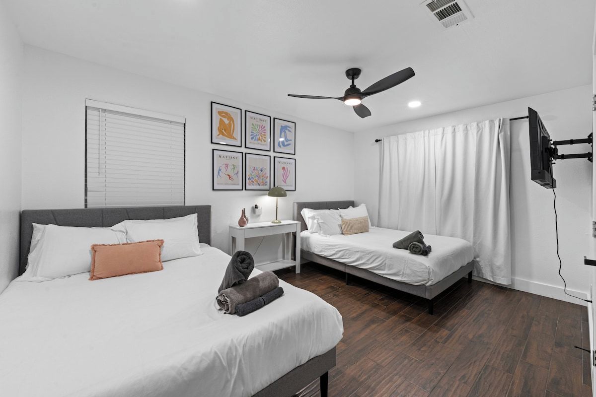 A hotel room with two beds, a ceiling fan, a TV, and artwork on the wall.