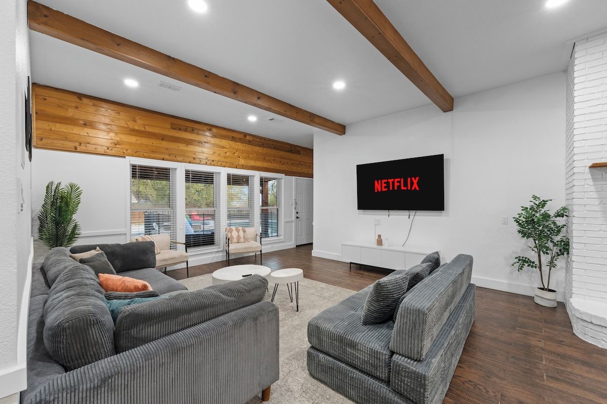 A modern living room with a large flat-screen TV mounted on the wall, a grey sectional sofa, a small white side table, and a potted plant.
