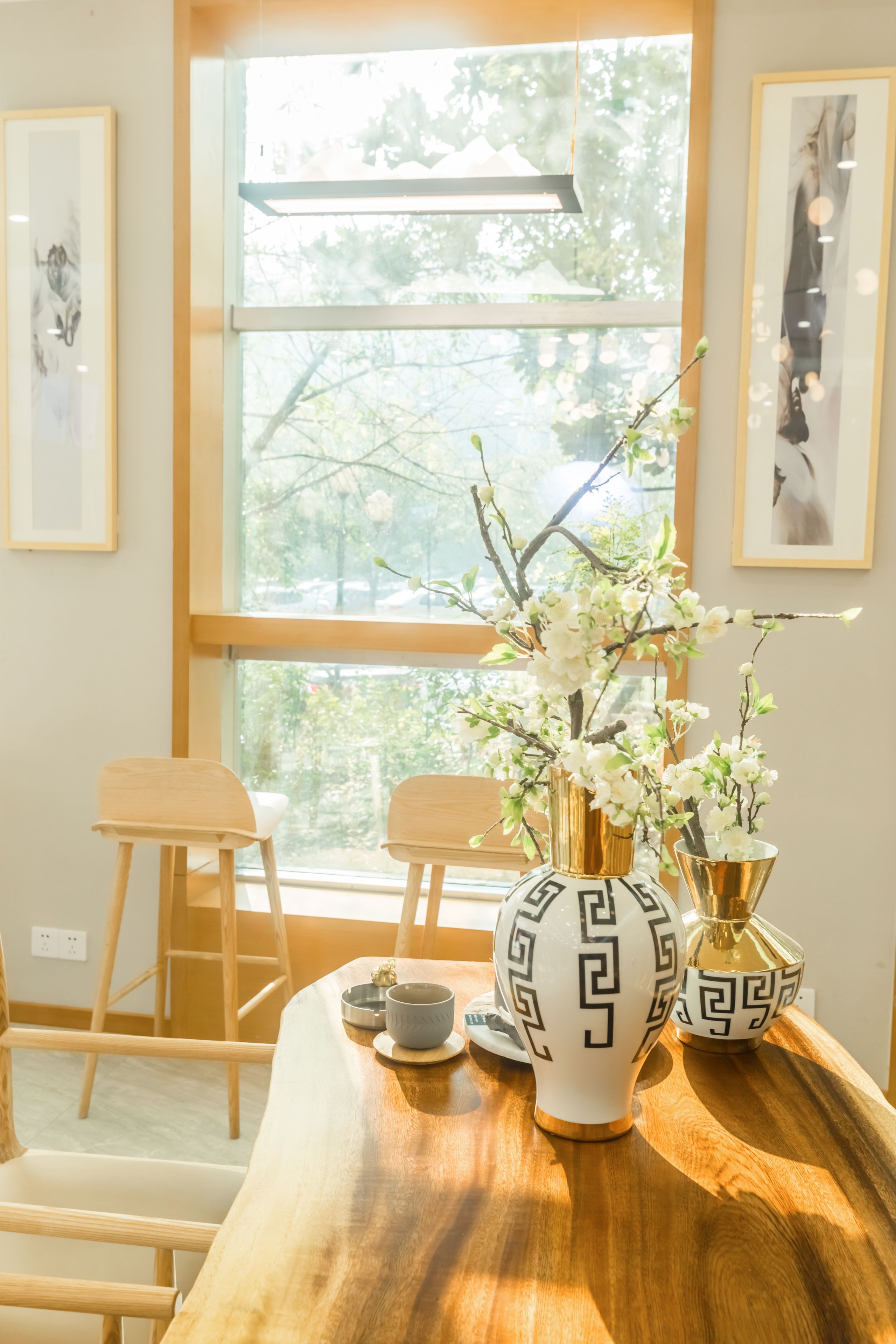 Una mesa de madera con un jarrón de flores, una taza y un platillo. Hay dos sillas y una escalera apoyada en la pared.