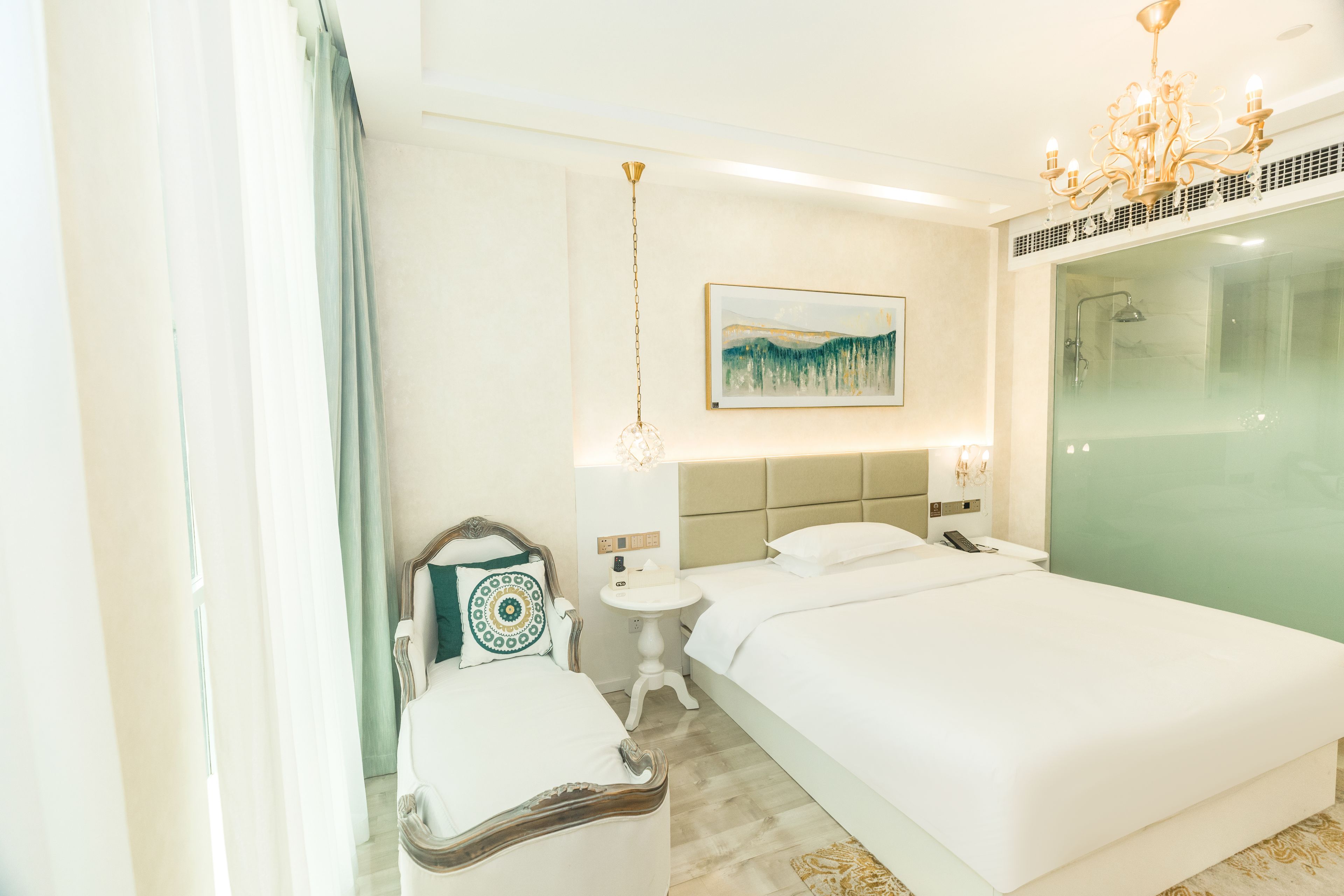 A hotel room with a bed, bedside table, lamp, artwork, and a shower area.