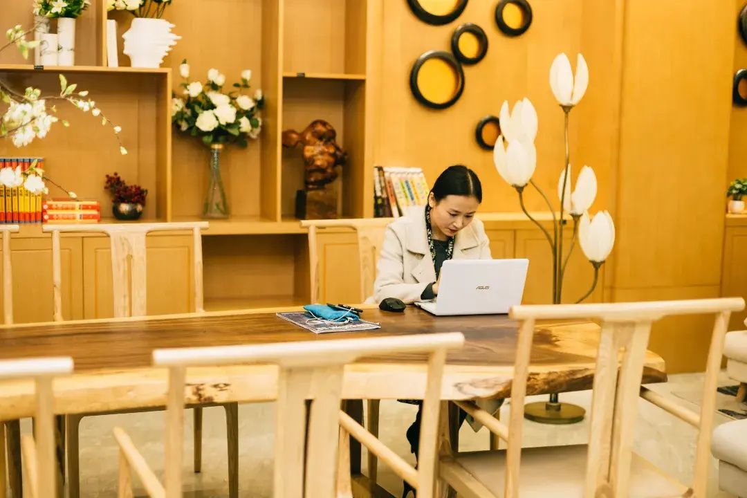 A person working at a wooden table with a laptop, surrounded by books and decorative items in a warmly lit room.