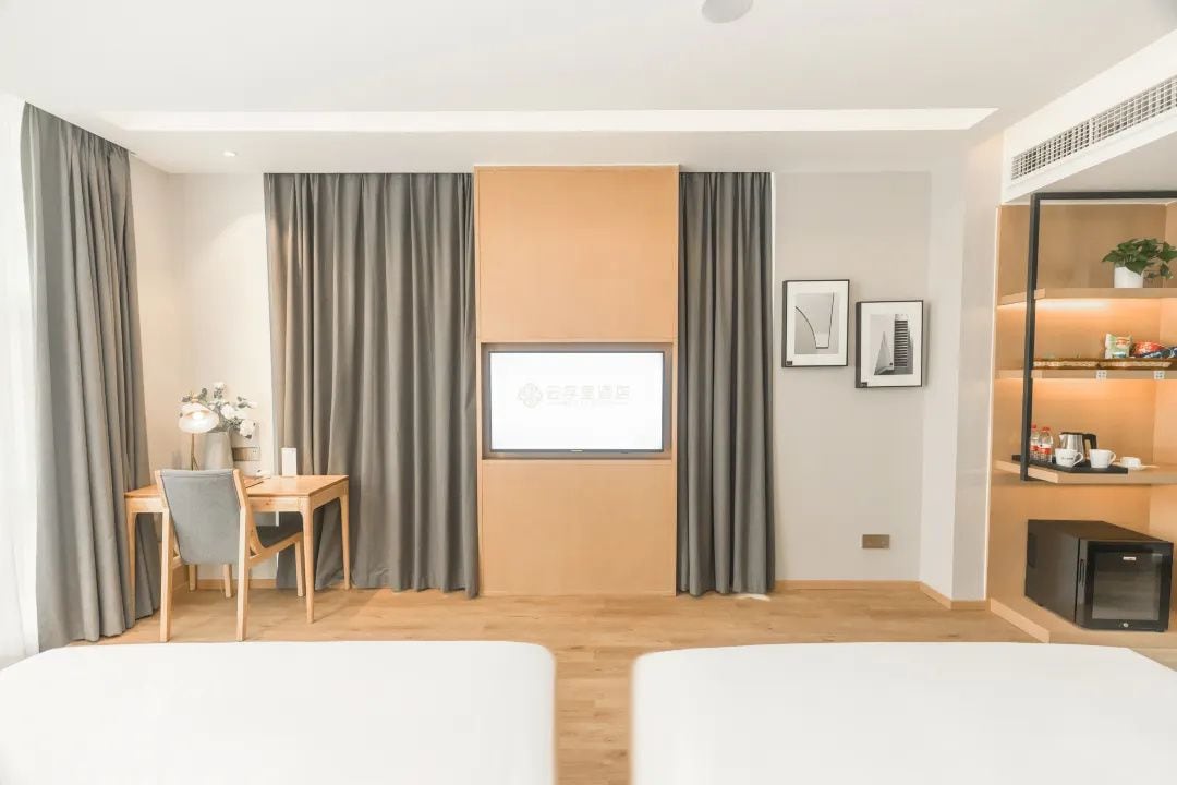 Habitación de hotel con dos camas, un escritorio, una silla, un refrigerador pequeño y una televisión en la pared.