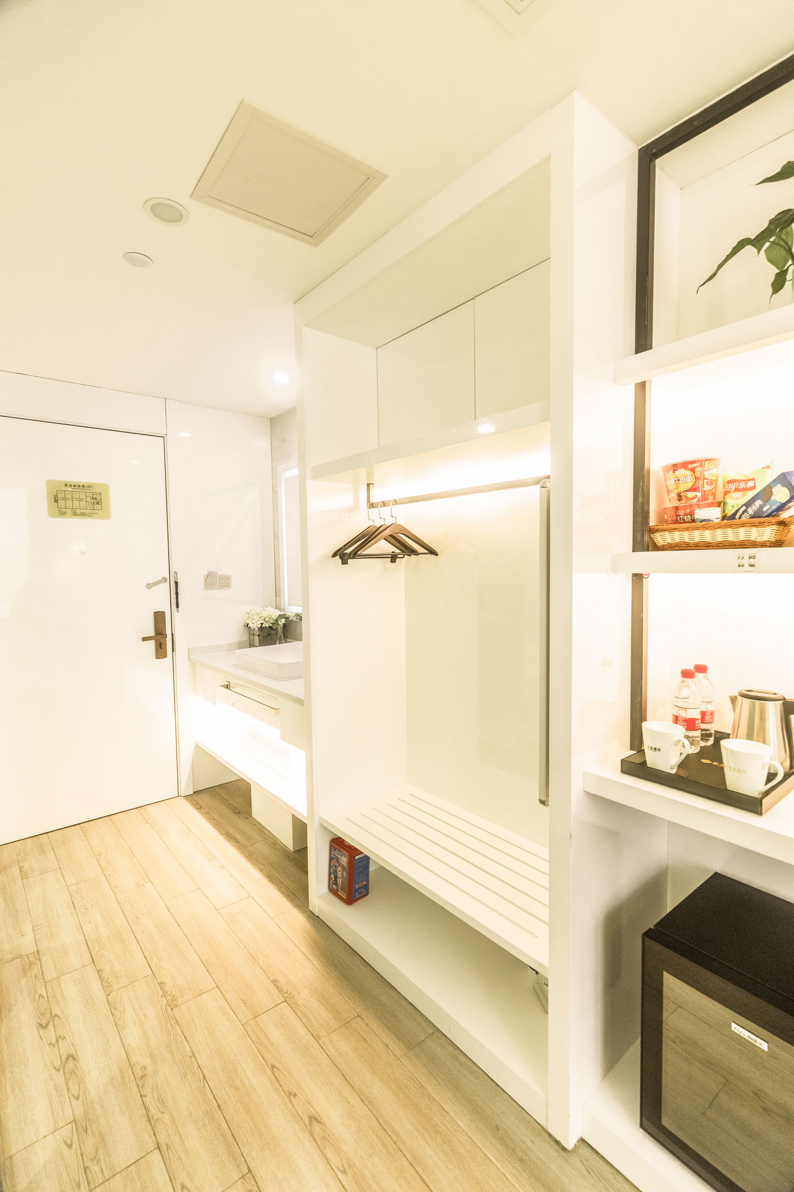Habitación de hotel con piso de madera, un armario blanco, una mesita de noche con lámpara y un estante con artículos de aseo.