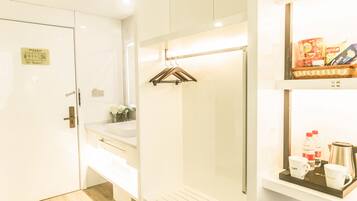 Habitación de hotel con piso de madera, un armario blanco, una mesita de noche con lámpara y un estante con artículos de aseo.