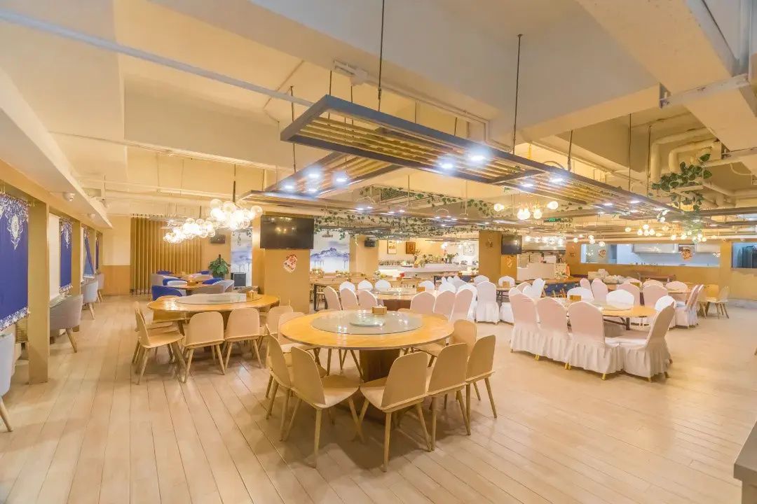 A spacious dining area with multiple round tables and chairs, a high ceiling with exposed beams, and a modern chandelier.