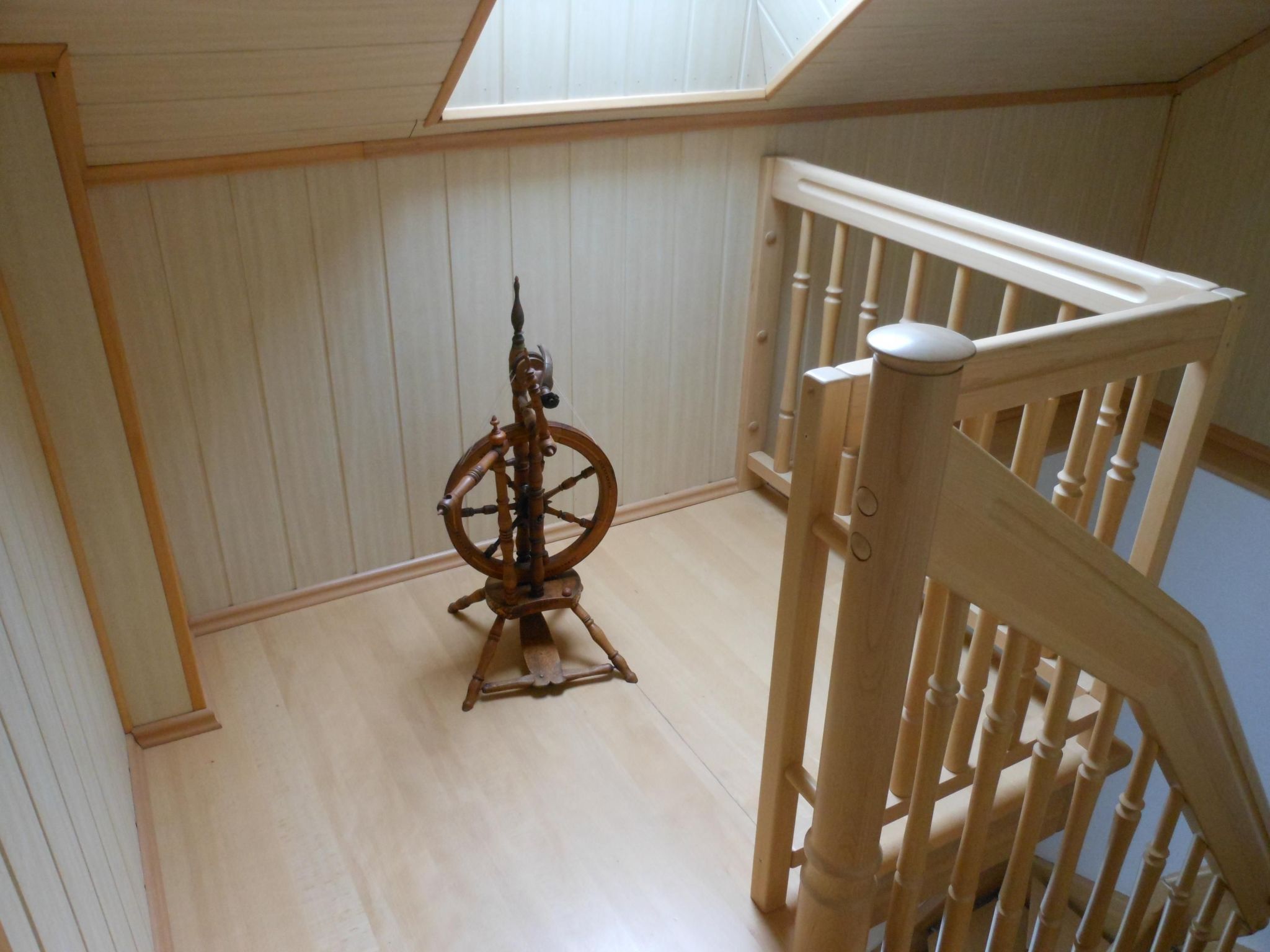 Un escalier en bois, doté d’une rampe, mène à une pièce dont le plafond est en pente et où se trouve un rouet.