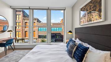 Habitación de hotel con una cama grande, un escritorio, una silla, una lámpara de escritorio, una lámpara de pie y vistas a la ciudad a través de una puerta corrediza de vidrio.