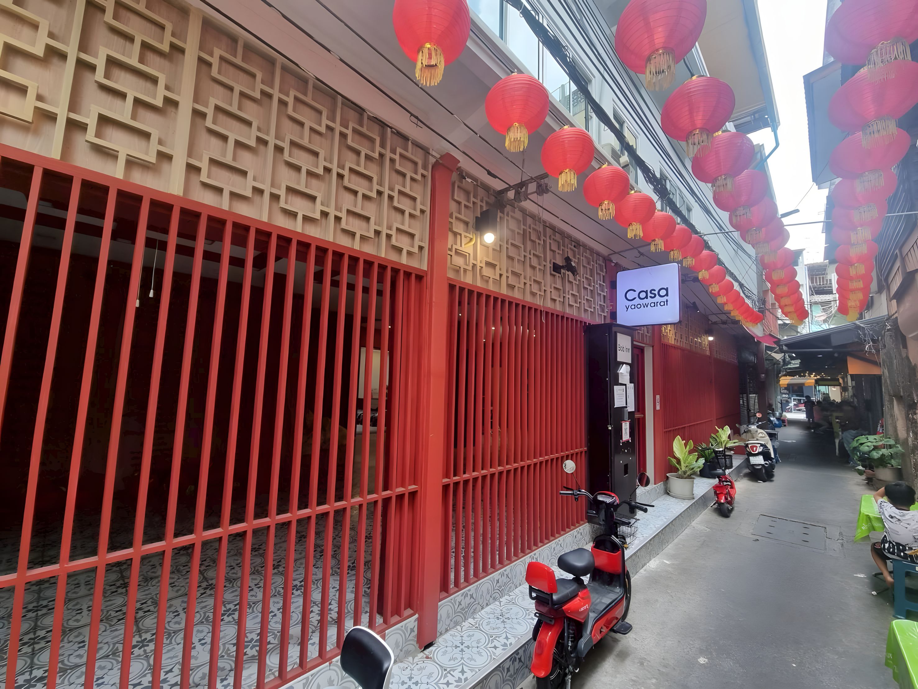 A narrow street with a red gate, a sign for 'Casa yaowarat,' and a scooter parked on the side.