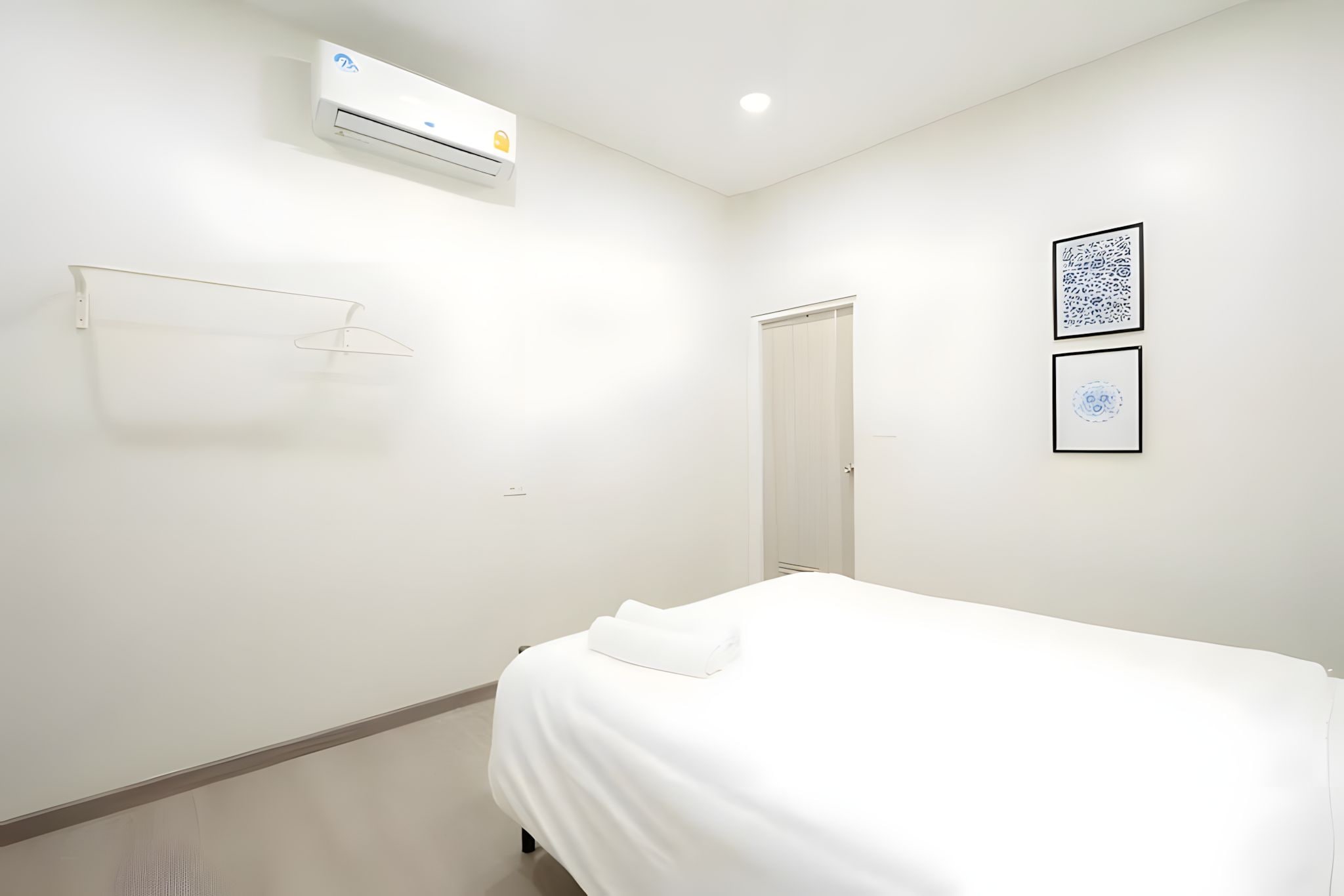 A minimalist hotel room with a bed, a wall-mounted air conditioner, a door, and two framed artworks.