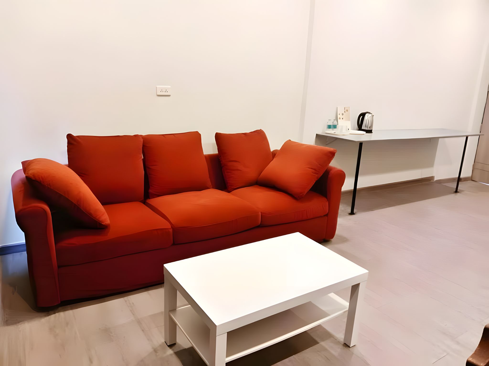 A modern living room with a red sofa, a white coffee table, and a minimalist white table with a kettle and a water bottle.