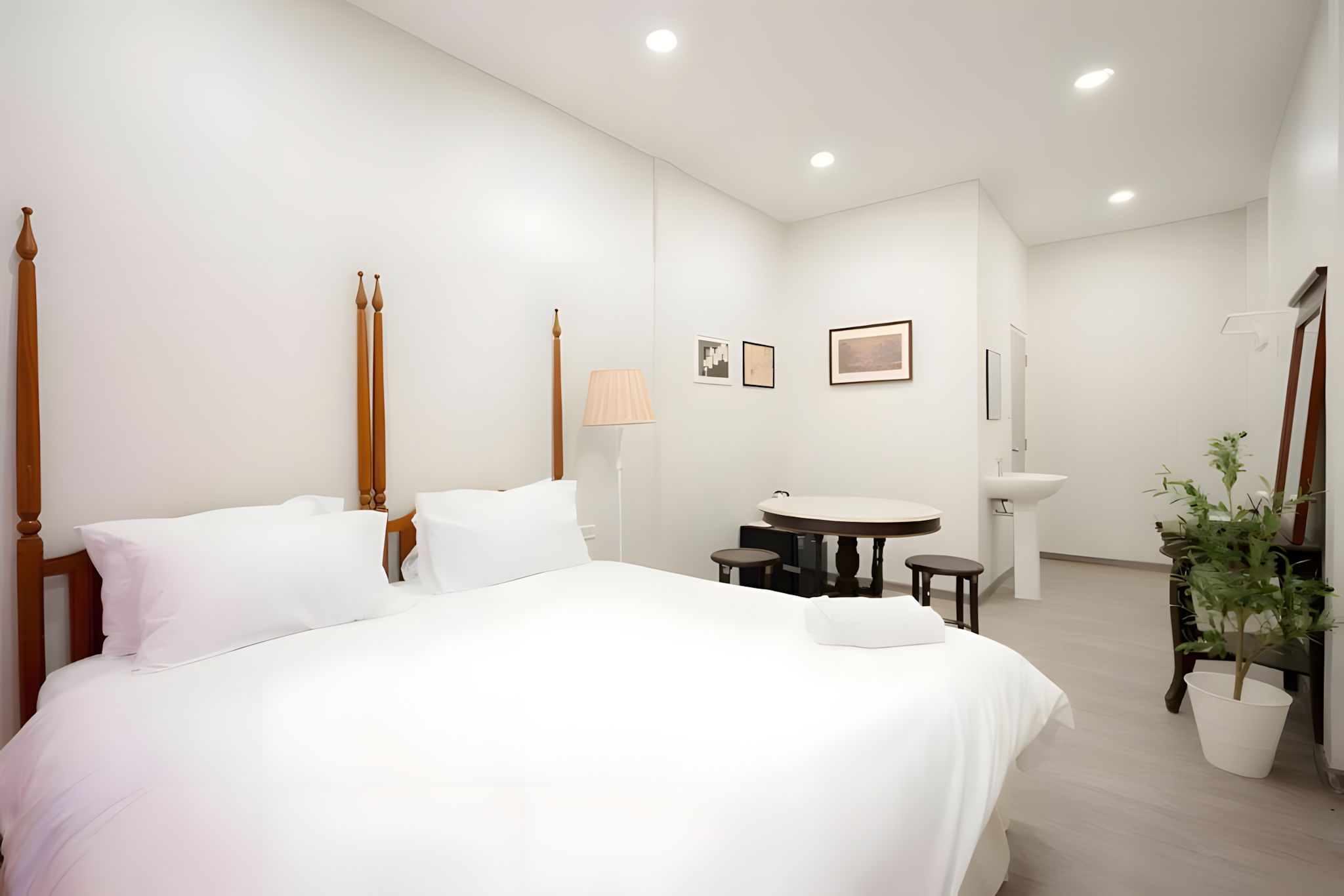 A hotel room with a large bed, bedside tables, a round table, a chair, a lamp, a sink, and a potted plant.