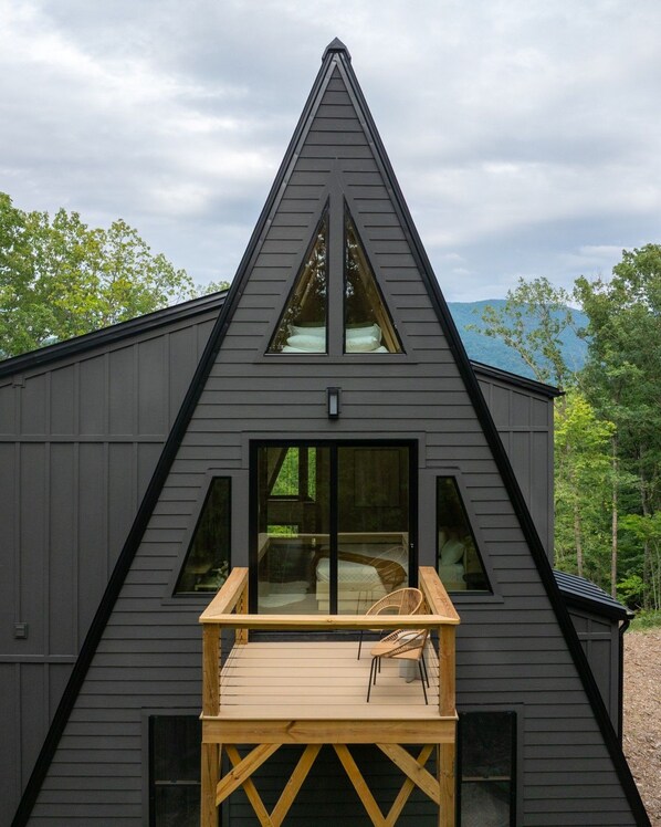 A modern house with a triangular roof, large windows, and a wooden deck with a chair. - Solhaus Shenandoah A-Frame | Park Views, Hot Tub, Theater & Game Room (Luray)