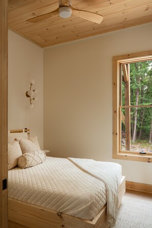 Una camera da letto con una ventola a soffitto in legno, un letto con materasso trapuntato, una lampada da comodino e una finestra vista alberi.