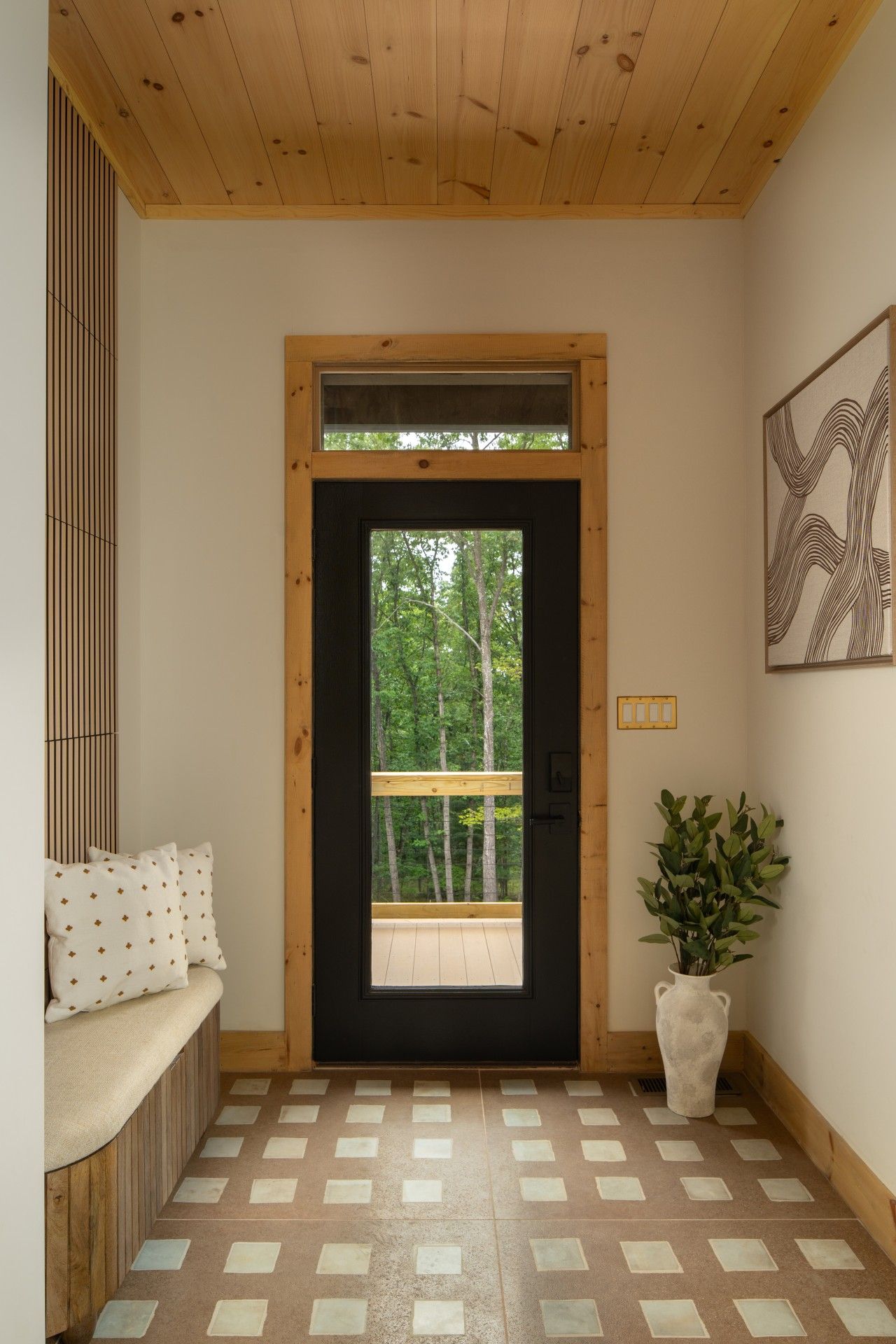 Una puerta de madera con vista a árboles, un banco con cojines estampados y una planta en maceta.