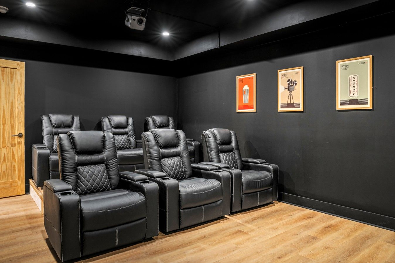 Un cine en casa con sillones reclinables de cuero negro, una puerta de madera y carteles de películas enmarcados en la pared.