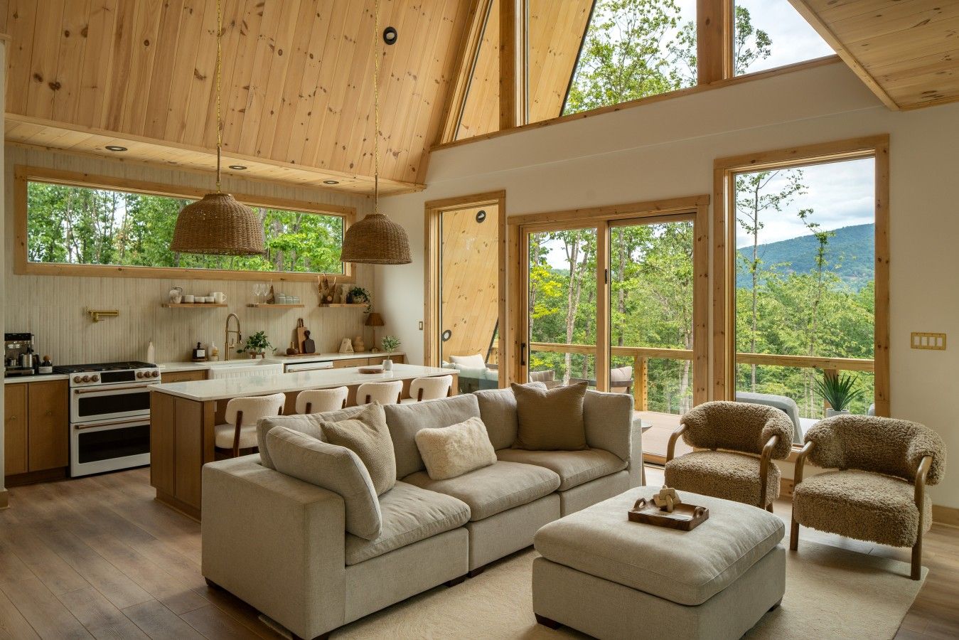Un vaste espace de vie comprenant un grand canapé, un pouf et deux fauteuils, une cuisine avec des meubles en bois et une cuisinière, ainsi que de grandes fenêtres offrant une vue sur l’extérieur.