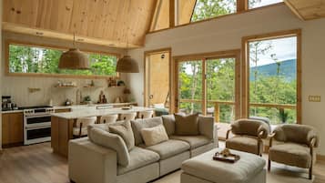 Un vaste espace de vie comprenant un grand canapé, un pouf et deux fauteuils, une cuisine avec des meubles en bois et une cuisinière, ainsi que de grandes fenêtres offrant une vue sur l’extérieur.