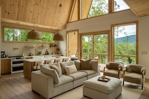 Ein großzügiger Wohnbereich mit einer großen Couch, einem Hocker und zwei Sesseln, eine Küche mit Holzschränken und einem Herd sowie große Fenster mit Blick ins Freie.