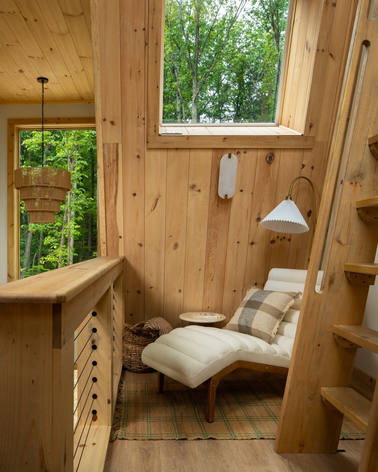 Interno di una baita di legno con un letto, una lampada a sospensione e una finestra vista foresta.