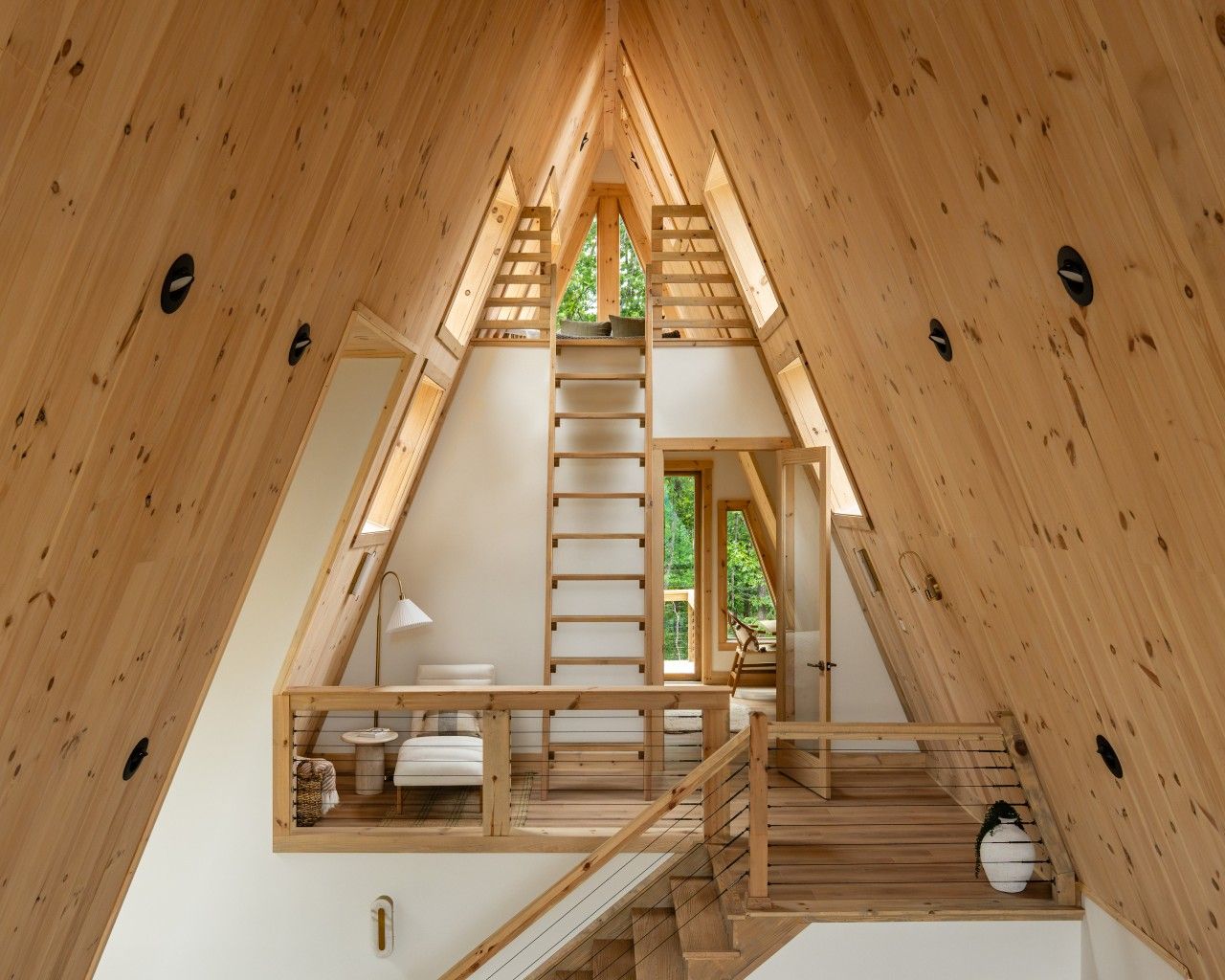 Un intérieur en bois avec une chambre à coucher située en mezzanine, un escalier et une vue sur la verdure environnante.