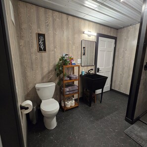 A bathroom with a wooden wall, a toilet, a sink, a shelf with towels, and a mirror.