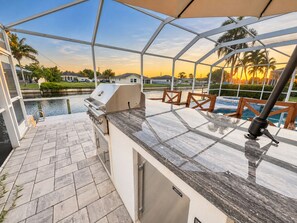 A covered outdoor patio with a grill, pool, and seating area. - Wonderful water view property with stunning boat Pool and built in Grill. (Cape Coral)