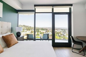Ein Hotelzimmer mit einem großen Bett, einem Schreibtisch mit Stuhl, einem Esstisch und Blick auf eine Stadtansicht durch große Fenster.