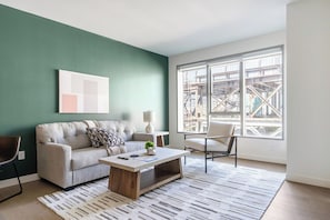 A living room with a sofa, coffee table, and a chair. There's a window with a view of construction outside. - Landing | Beautiful 1BD, Rooftop Lounge, Gym (Los Angeles)