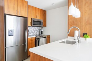 A modern kitchen with stainless steel appliances, wooden cabinets, a white countertop, and pendant lighting. - Landing | Incredible 1BD, Gym, Rooftop Lounge (Los Angeles)