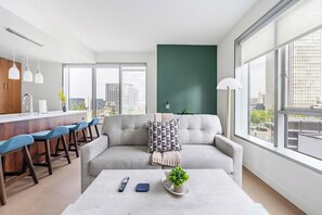A modern living room with a grey sofa, a coffee table with remote controls, and a view of the city through large windows. - Landing | Spectacular 2BD, Gym, Rooftop Lounge (Los Angeles)
