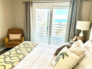 Habitació d'hotel amb un llit, una cadira, una làmpada i vista a la platja per la finestra.