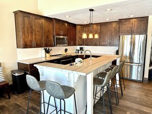 A modern kitchen with wooden cabinets, a stainless steel refrigerator, and a central island with bar stools. - Your Ultimate Bear Lake Lakeside Escape! (Garden City)