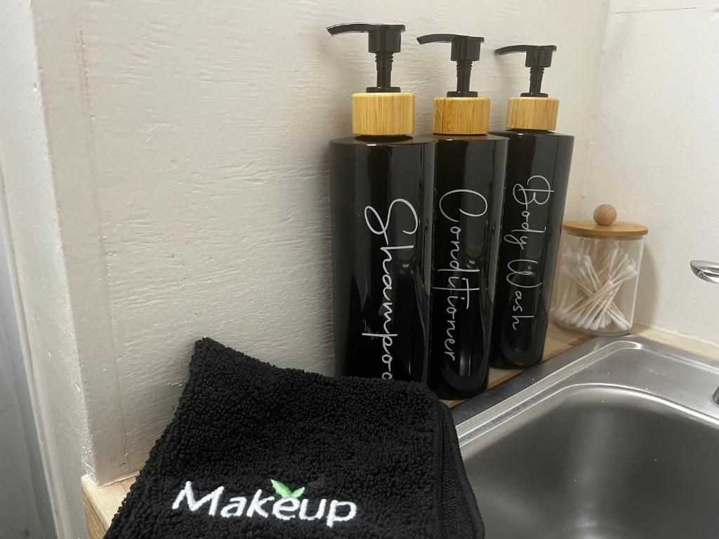 Un baño con una toalla negra en el suelo, un lavabo y tres botellas negras con dispensador, etiquetadas como 'Body Wash' y 'Shampoo'.