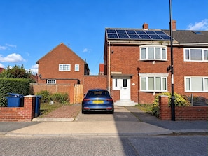 Een huis van baksteen met twee verdiepingen, een auto op de oprit, zonnepanelen op het dak en een brievenbus op de veranda.
