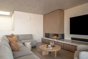 A modern living room with a gray sofa, a round coffee table, a fireplace, and a large flat-screen TV mounted on the wall. - Equinox | Ocean Front, Heated Pool, Gym, Sauna (Gerroa)