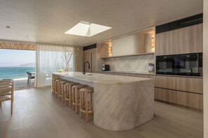 A modern kitchen with a central island, bar stools, and a view of the sea. - Equinox | Ocean Front, Heated Pool, Gym, Sauna (Gerroa)