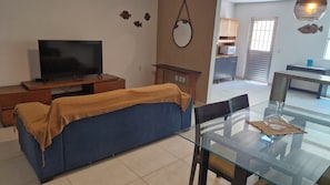 A living room with a sofa, a glass dining table, a TV, and a kitchen area in the background. - Casa Canopus is 1 minute from the beach, then enjoy the pool. Relax and have fun. (Japaratinga)