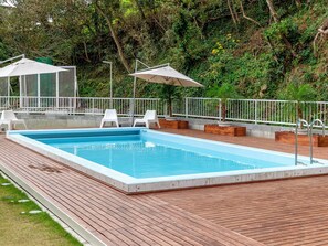A rectangular swimming pool with a wooden deck, surrounded by white chairs and umbrellas, set against a backdrop of dense greenery.