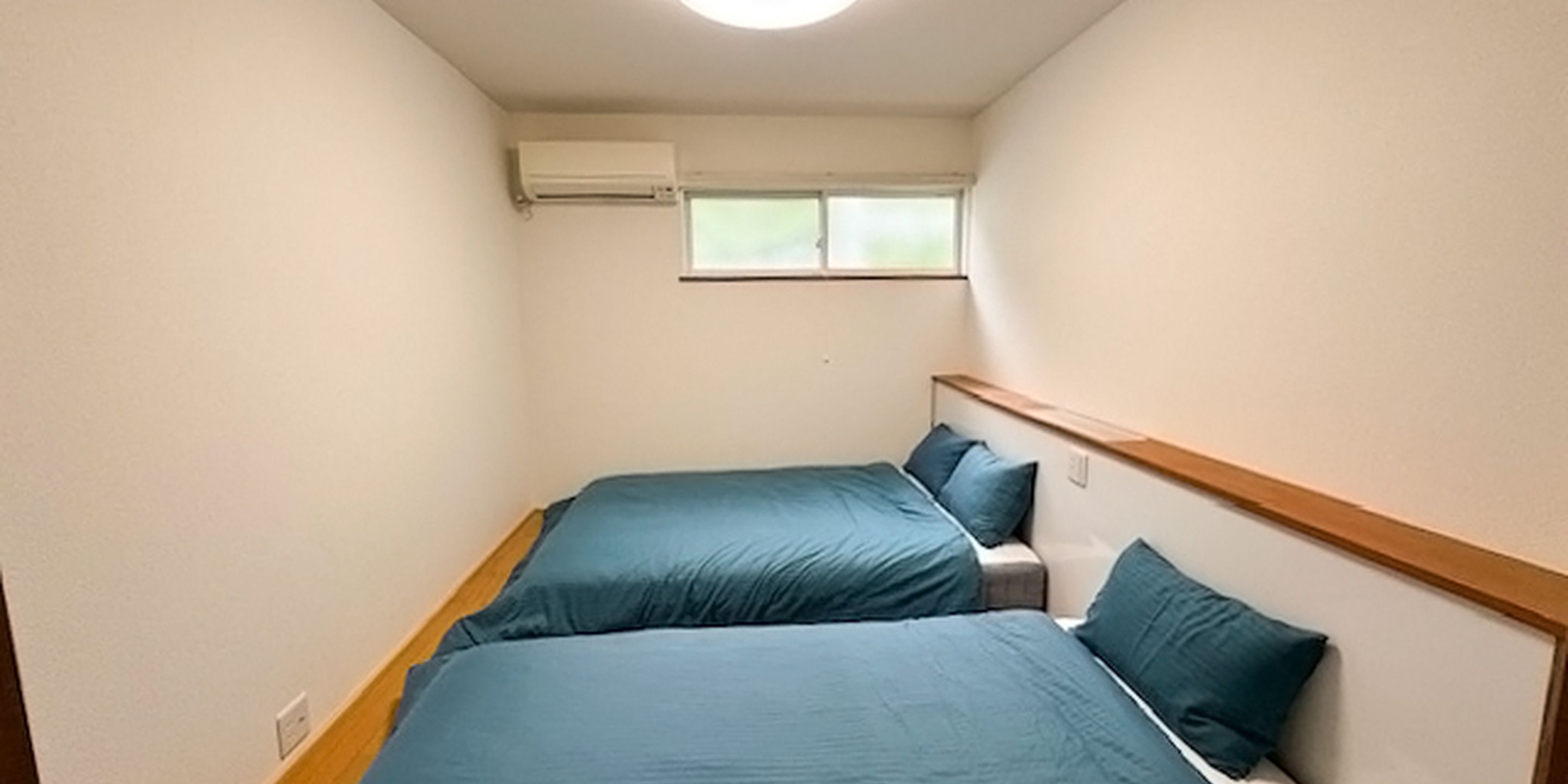 Quarto pequeno e estreito com duas camas, uma janela e aparelho de ar condicionado.