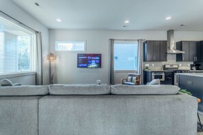 Una sala moderna con un sofá seccional gris, un televisor de pantalla plana montado en la pared y una cocina con armarios oscuros y electrodomésticos de acero inoxidable.