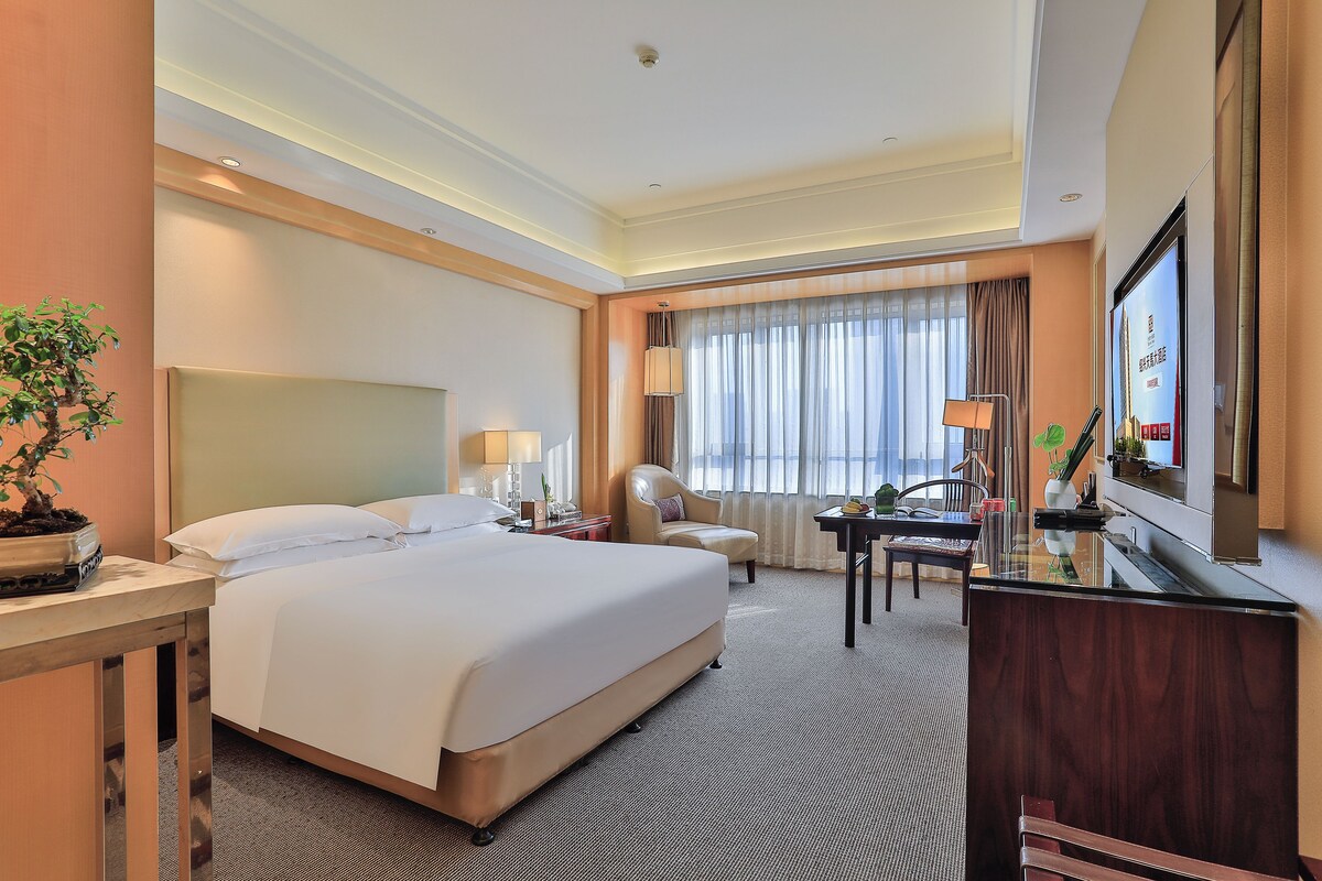 A hotel room with a large bed, a desk with a lamp, a chair, a television, and a potted plant.