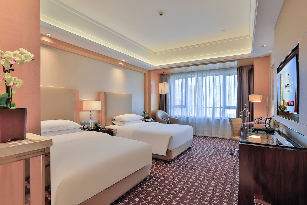A hotel room with two beds, a desk with a telephone, a mirror, a TV, and a window with curtains.