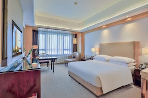 A hotel room with a large bed, a desk with a chair, a dining table, and a view of the city through the window.