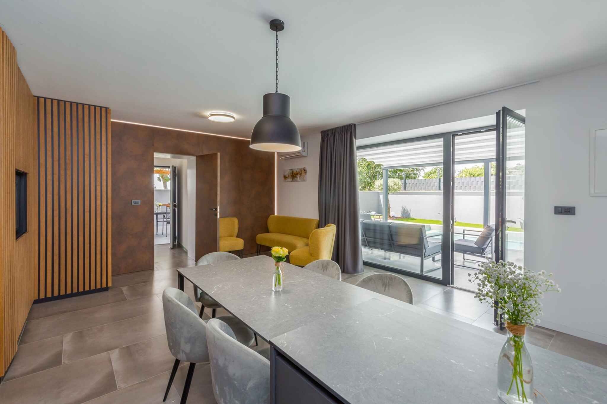 A modern kitchen with a central island, dining chairs, a hanging pendant light, and a view through large windows to an outdoor area.