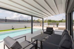 Ein moderner Außenbereich mit Pool, einer überdachten Terrasse und Blick auf ein Sportfeld.