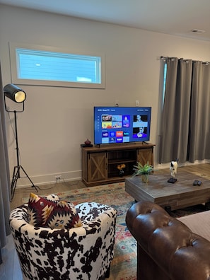 A living room with a flat-screen TV mounted on a wooden stand, a floor lamp, a patterned ottoman, a leather sofa, and a decorative skull on a coffee table. - Spacious 4-bedroom house across from Pickleball Park near Uptown (Charlotte)