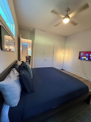 Un dormitorio con cama, ventilador de techo, televisor montado en la pared y puerta que da a otra habitación.