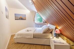 Ein Schlafzimmer mit Bett, Nachttisch mit Lampe, Fenster mit Blick auf Bäume und einem Bild an der Wand.