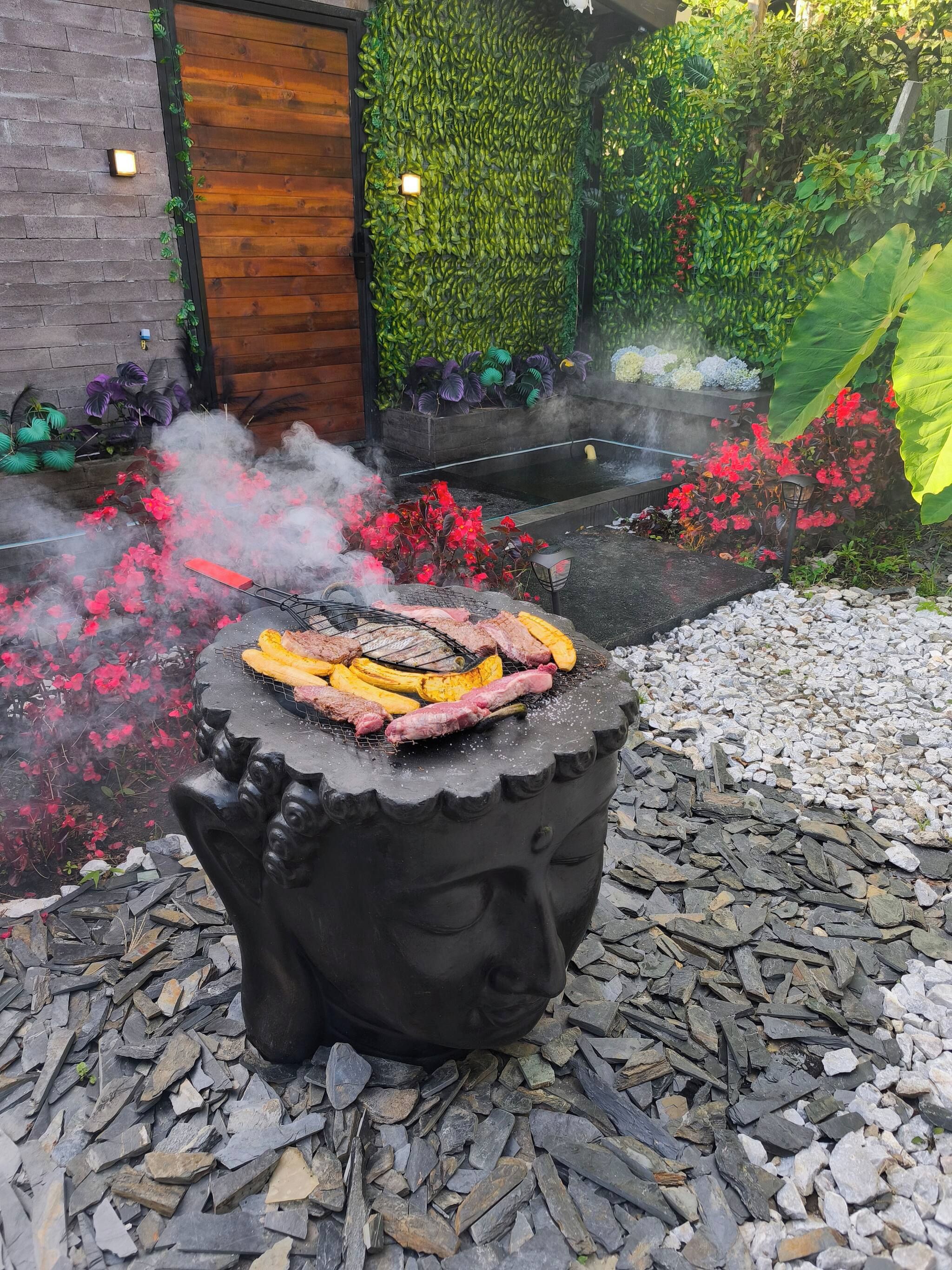 Un barbecue extérieur où l’on grille différentes sortes de viandes et de légumes, le tout entouré d’un jardin agrémenté de fleurs rouges et de verdure.