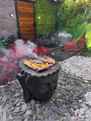 Un barbecue extérieur où l’on grille différentes sortes de viandes et de légumes, le tout entouré d’un jardin agrémenté de fleurs rouges et de verdure.